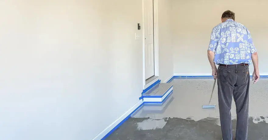 Mann beschichtet den garagenboden mit einem Roller. Der Boden ist teilweise mit einer grauen Beschichtung bedeckt, die Ränder sind mit blauer Klebefolie abgeklebt.
