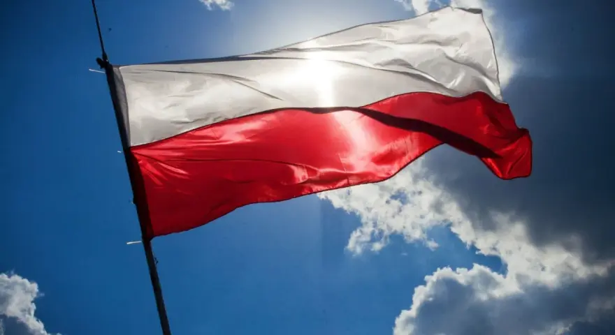 Biało-czerwona flaga Polski powiewa na tle błękitnego nieba z chmurami. Słońce przebija się przez materiał, podkreślając jego fakturę. Wymiary flagi polski są standardowe, proporcje zachowane.