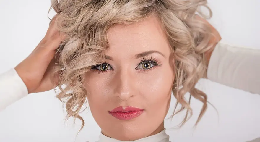 Blondynka z kręconymi włosami, z nonszalanckim gestem poprawiająca fryzurę. Jej oczy przyciągają uwagę, a usta są pomalowane na różowo.