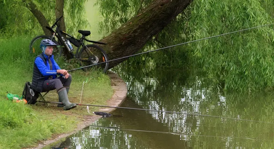 Wędkarstwo spławikowe na stawie. Mężczyzna w kasku i kamizelce siedzi na krześle, trzymając wędkę. Obok rower.