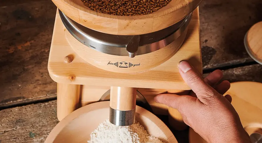 Holzgetreidemühle mit Getreide oben und frisch gemahlenem Mehl unten, Hand bedient die Mühle.