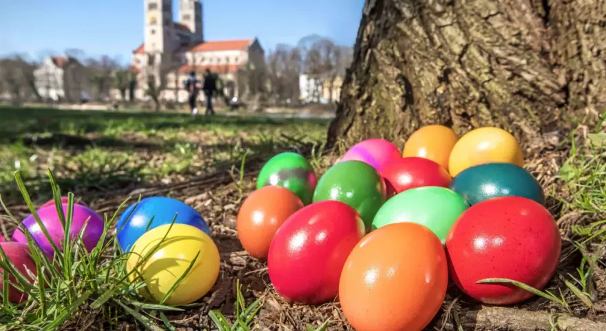 Bunte Ostereier liegen im Gras vor einem Baum. Im Hintergrund ist eine Kirche zu sehen. Die Suche nach den Eiern ist ein schöner Teil der Ostern Bedeutung.