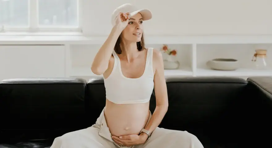 Kobieta w ciąży, czując uderzenia gorąca, poprawia czapkę. Siedzi na czarnej kanapie w jasnym wnętrzu.