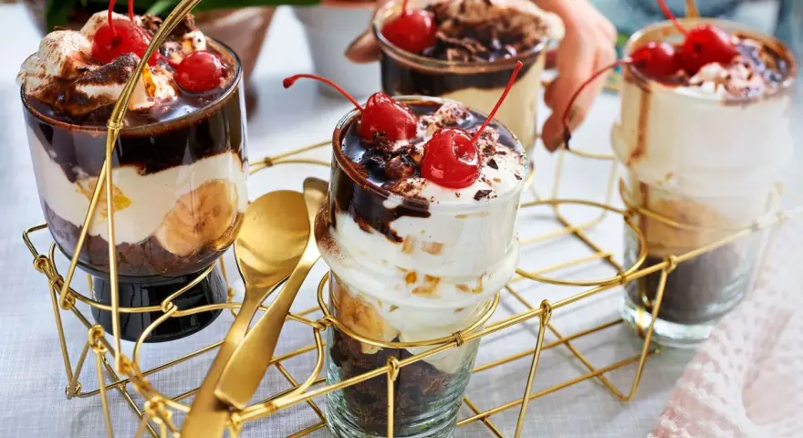 Schokoladige Dessert-Rezepte in Gläsern mit Bananen, Sahne und Kirschen. Goldene Löffel liegen bereit.
