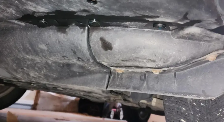 Underneath a car, dark fluid is leaking from a seam. A small puddle of blue fluid is visible, suggesting a coolant leak.