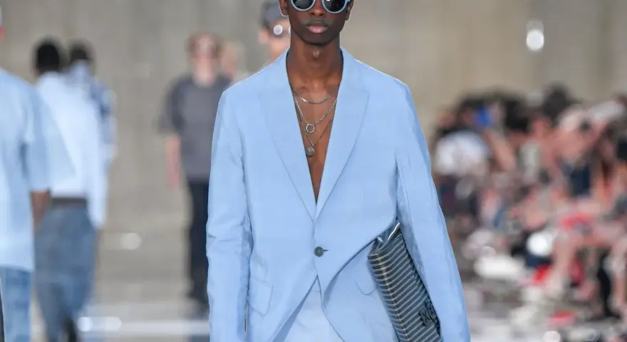 Modello con outfit uomo elegante senza giacca, completo azzurro, occhiali da sole blu e borsa a righe.