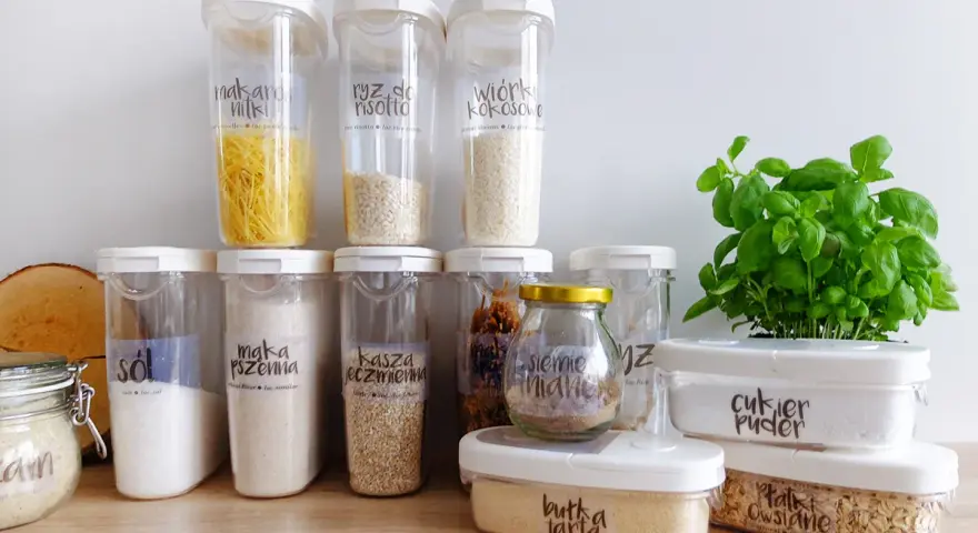 Pojemniki na żywność z etykietami pokazują, jak przechowywać pokrywki i produkty sypkie. Widać makaron, ryż, sól, mąkę, kaszę, cukier puder i płatki owsiane.
