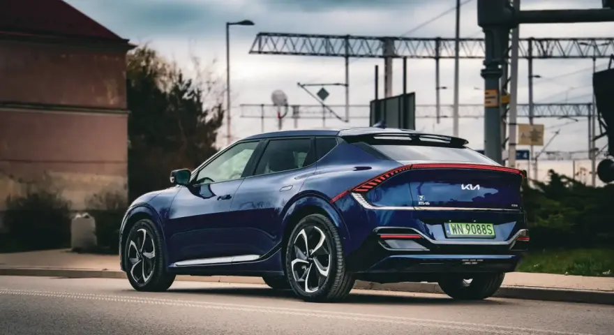 Granatowe Kia EV6 GT z charakterystycznym pasem świateł z tyłu, zaparkowane przy drodze z widoczną infrastrukturą kolejową.