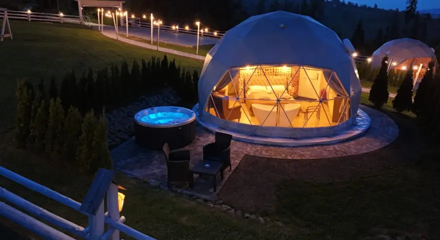 Nocny widok na luksusowy camping w Tatrach. Dwa kopułowe namioty, jeden z wanną i łóżkiem, drugi z oświetleniem. Obok jacuzzi i meble ogrodowe.