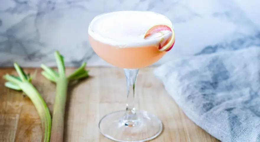 En rosa rabarber sour-drink med skum och en rabarberdekoration. Två stjälkar rabarber ligger bredvid.