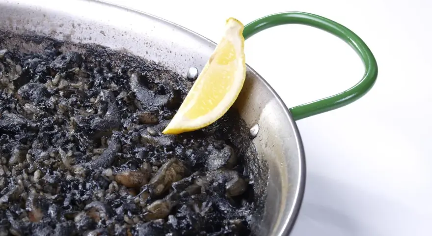 Arroz negro en paellera, con trozos de calamar y un gajo de limón.