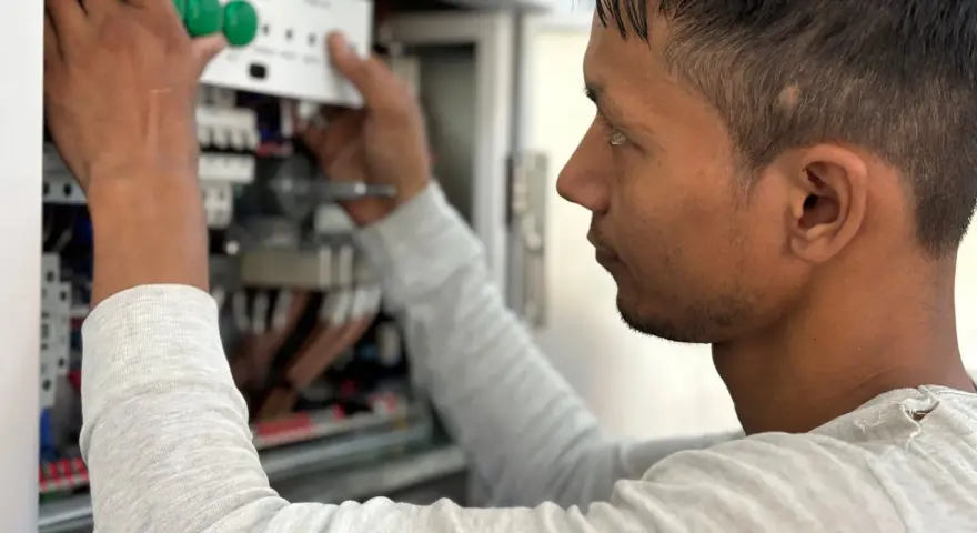 Mężczyzna w jasnej bluzie pracuje przy panelu elektrycznym, montując białe urządzenie z zielonymi przyciskami.