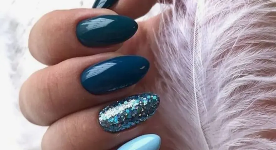 Unghie per l'inverno: unghie blu petrolio e azzurre con glitter scintillanti, accanto a una soffice piuma bianca.