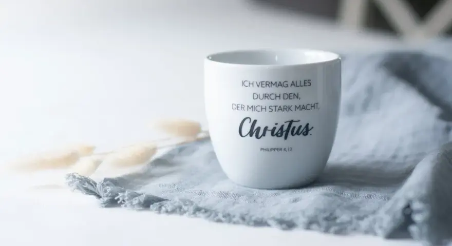 Weiße Tasse mit dem Schriftzug "Ich vermag alles durch den, der mich stark macht, Christus. Philipper 4,13" auf einem grauen Stoff.