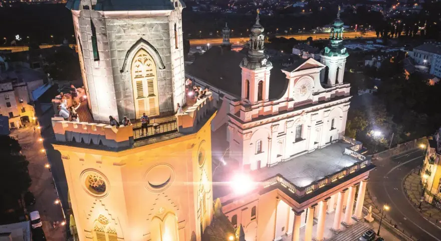 Wieża trynitarska nocą, z ludźmi na balkonie i oświetloną fasadą kościoła.