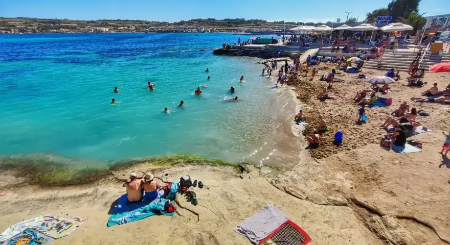 Słoneczna plaża na Malcie w lutym. Ludzie kąpią się w turkusowej wodzie, opalają na piasku i odpoczywają pod parasolami.