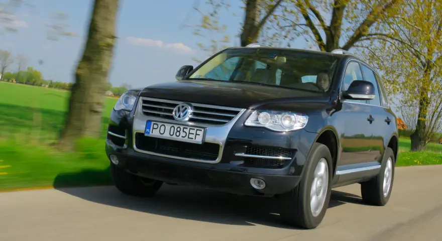 Czarny VW Touareg 3.0 TDI w ruchu, z widocznymi wadami lakieru i drobnymi niedoskonałościami, na tle zielonych pól i drzew.