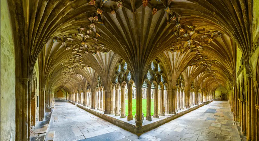 A grand cloister with vaulted ceilings, reminiscent of a monastery floor plan. Sunlight streams through arched windows onto the stone floor.