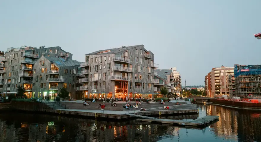 Moderna byggnader längs en kanal i Oslo. Folk sitter på trappor vid vattnet. Besök Oslo för en härlig kväll.