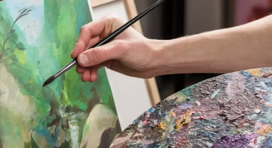 Main d'artiste peignant une scène verdoyante avec une peinture à l'acrylique. Palette de couleurs vibrantes.