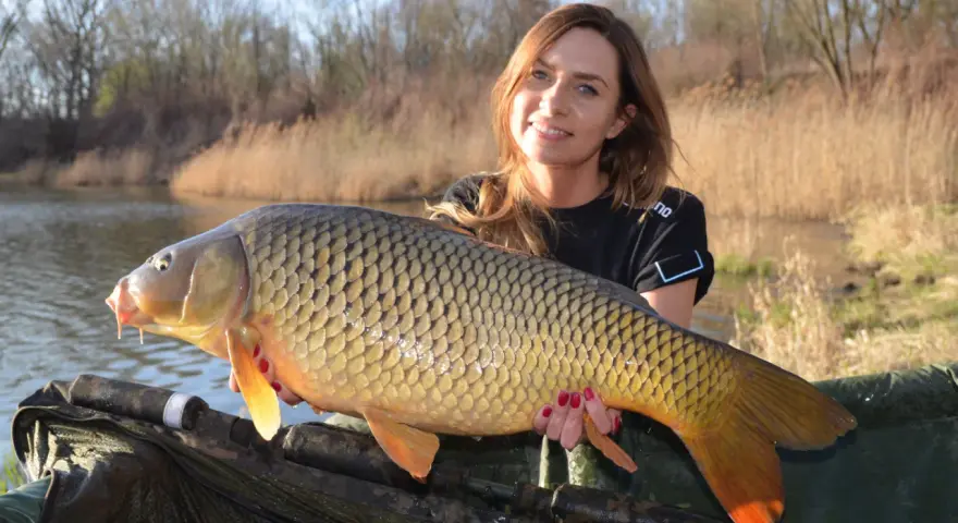 Uśmiechnięta wędkarz trzyma ogromnego karpia, pokazując, na co biorą karpie w stawie.