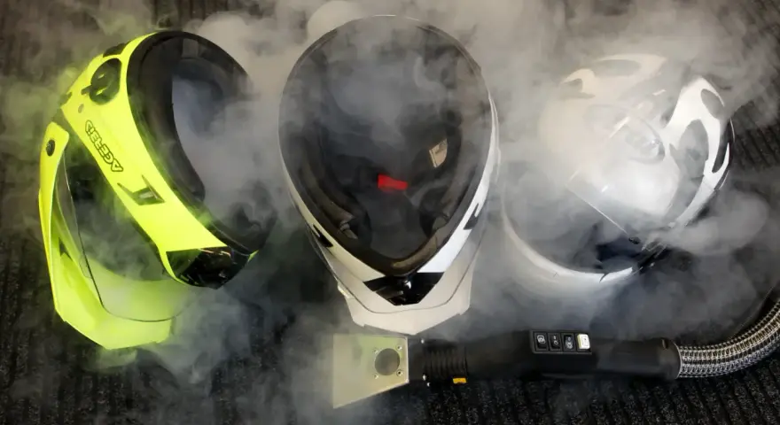 Pulizia del casco con vapore. Tre caschi, uno giallo fluo, uno bianco e nero, uno bianco e nero con visiera, circondati da fumo.
