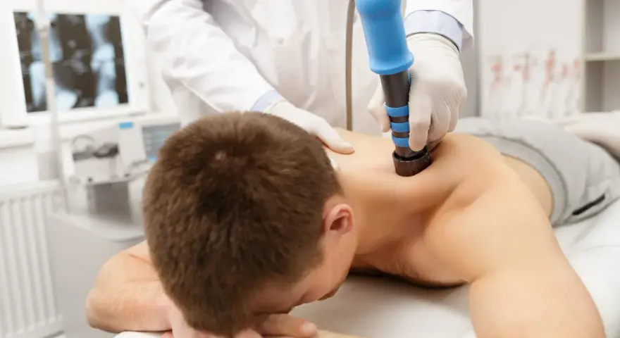 Arzt behandelt HWS-Syndrom mit Stoßwellentherapie. Ein Mann liegt auf dem Bauch, während ein Arzt mit einem blauen Gerät auf seinen Rücken drückt.
