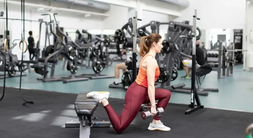 Kvinna utför bulgarian split squat med hantlar på gym.