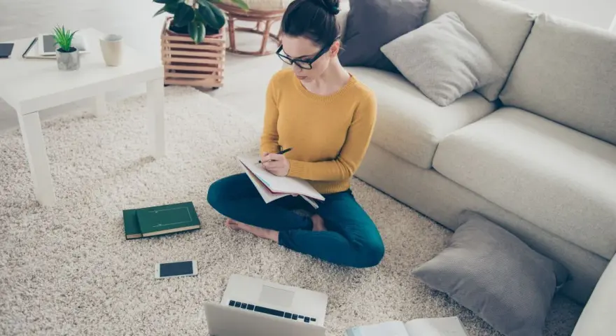 Dziewczyna w okularach uczy się w domu, przygotowując się do egzaminów licealnych. Na dywanie ma laptopa, książki i notatnik.