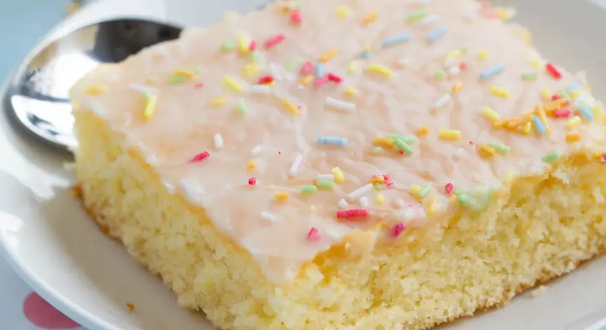 Ein Stück saftiger Fantakuchen vom Blech, mit Zuckerguss und bunten Streuseln bestreut, auf einem Teller mit Löffel.