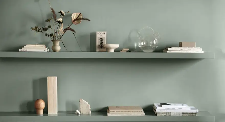 Två gröna hyllplan med böcker, vas med blommor, glasburk och dekorativa föremål. Enkel och stilren inredning.