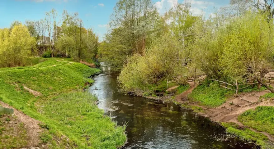 Ein idyllischer Panke Wanderweg schlängelt sich durch üppiges Grün. Ein kleiner Fluss fließt friedlich, gesäumt von Bäumen und Wiesen.
