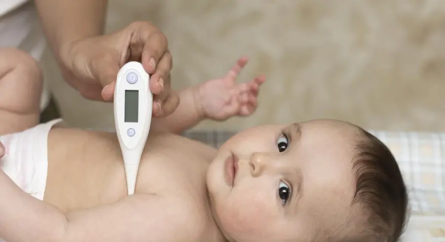 Dłoń trzyma termometr, mierząc temperaturę noworodka. Dziecko leży na plecach, patrząc w górę.