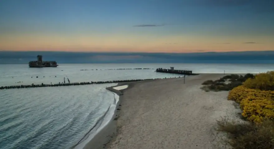 Zachód słońca nad morzem w Gdyni. Na wodzie widać betonową fortecę, a na brzegu piaszczystą plażę i babie doły.