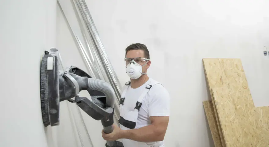 Operaio con maschera e occhiali sta per scartavetrare muro con una levigatrice.