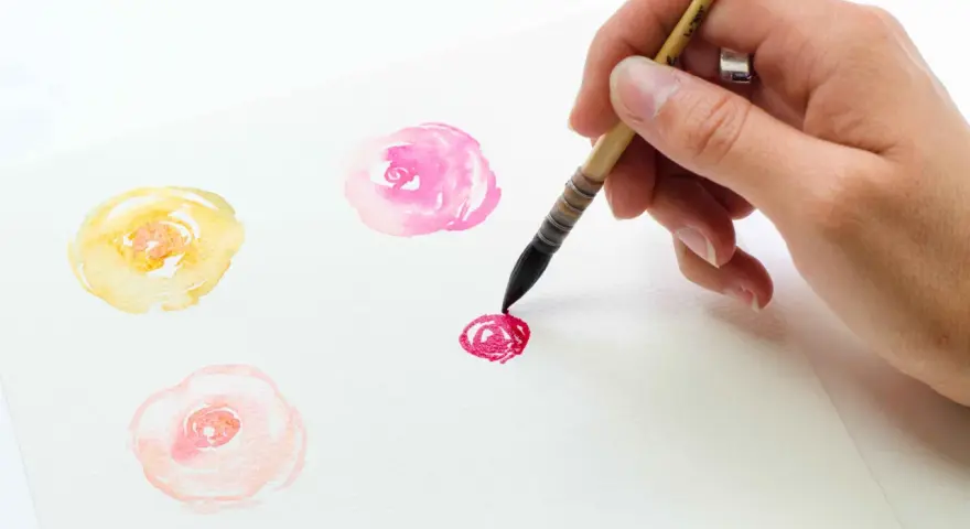 Un dessin de champs de fleurs facile à l'aquarelle. Une main peint des roses délicates en jaune, rose et rouge sur du papier blanc.