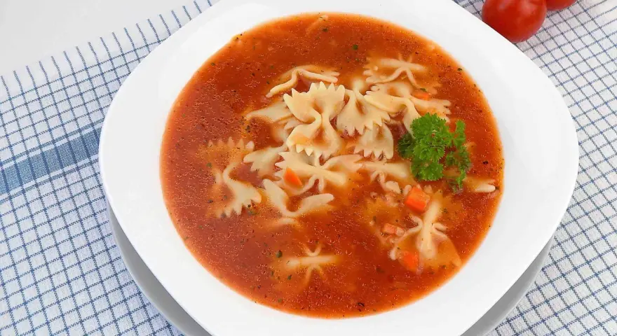 Zupa pomidorowa z makaronem farfalle, marchewką i natką pietruszki na kratkowanym obrusie.