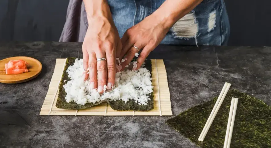Ręce rozkładają ryż na nori, przygotowując się do zwinięcia sushi. Obok leżą pałeczki i talerzyk z imbirem, idealny dodatek do sushi.