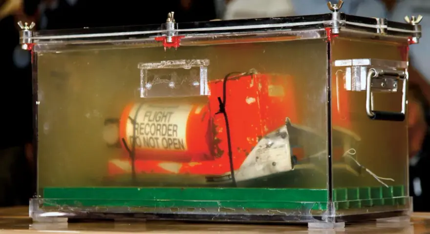 Pomarańczowa skrzynka "czarnej skrzynki" z napisem "FLIGHT RECORDER DO NOT OPEN" w przezroczystej obudowie, dowód w sprawie wypadku lotniczego.