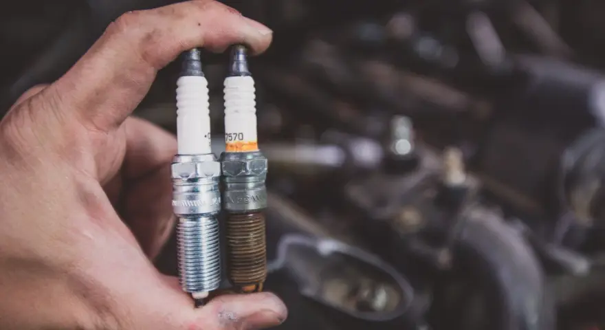 Dłoń trzyma dwie świece zapłonowe: jedną nową, błyszczącą, a drugą zużytą, z brązowym nalotem. Widać, jak wygląda zużyta świeca zapłonowa.