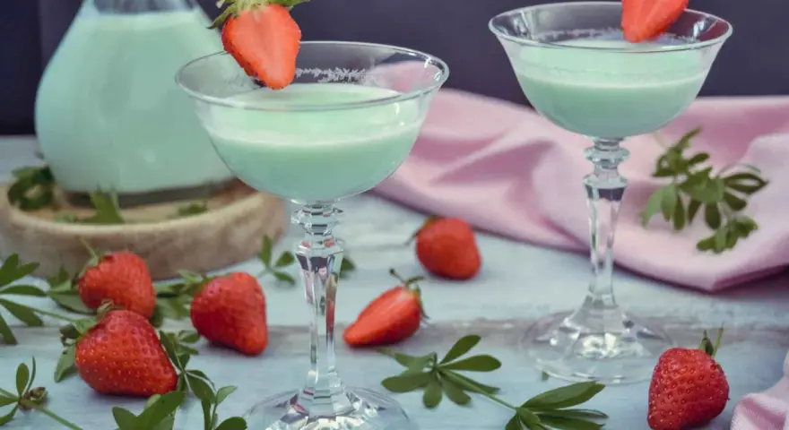 Zwei Gläser mit hellgrünem Likör, garniert mit Erdbeeren. Ein Krug mit dem Likör und frische Erdbeeren deuten darauf hin, dass man Waldmeister Schnaps selber machen kann.