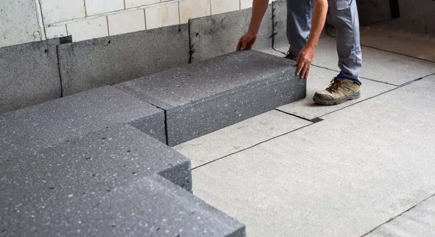 Pracownik układa szary styropian pod podłogówkę. Elementy tworzą izolację na betonowym podłożu.