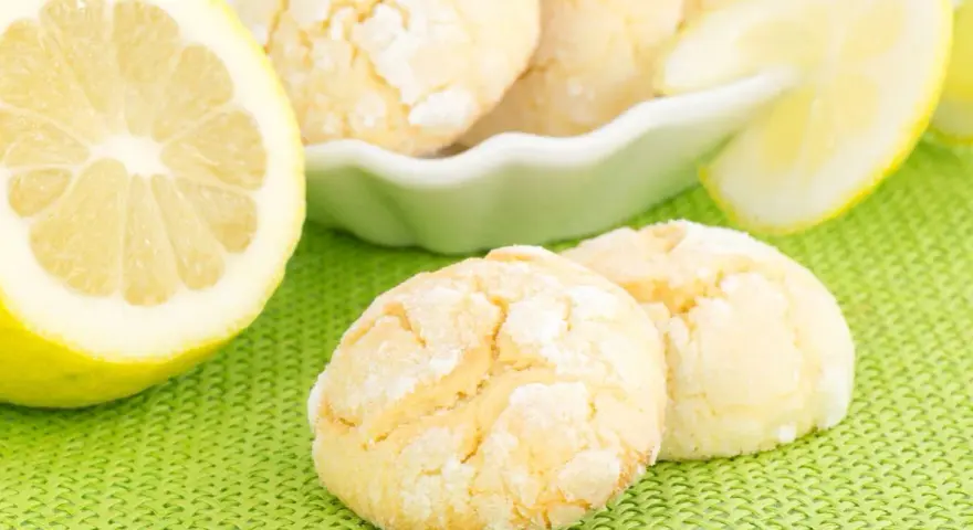 Biscotti al limone spolverati di zucchero a velo, accanto a fette di limone fresco su sfondo verde.