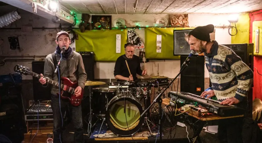 Un groupe de musique punk des années 90 joue dans un local sombre. Un guitariste, un batteur et un claviériste sont sur scène.