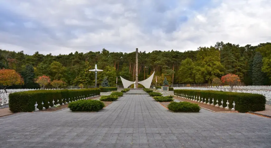 Uroczysty cmentarz wojenny z białymi krzyżami i pomnikiem pośrodku. W tle las, jesienne drzewa dodają koloru.