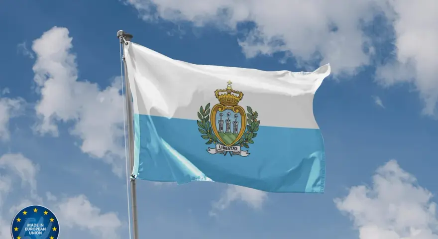 Flaga San Marino powiewa na tle błękitnego nieba z chmurami. Herb z trzema wieżami i koroną zdobi biało-błękitny materiał.