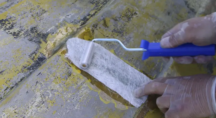 Händer med handskar arbetar med glasfibermatta och en roller för att laga en skadad yta.