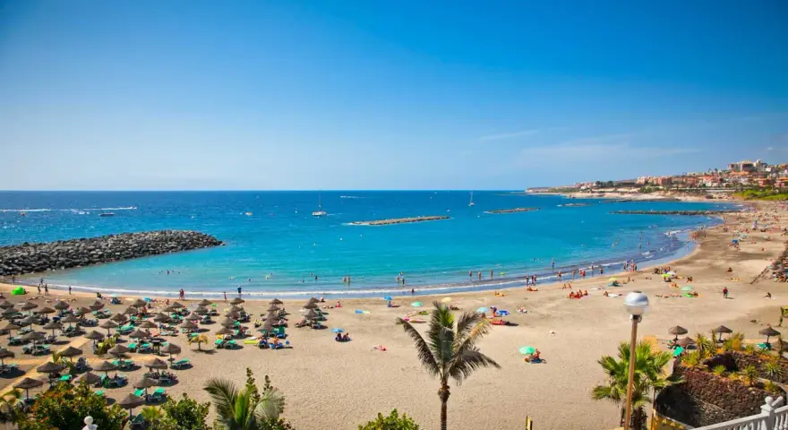 Spiaggia affollata con ombrelloni e lettini, mare azzurro e città costiera sullo sfondo. Sembra l'isola più bella delle Canarie.