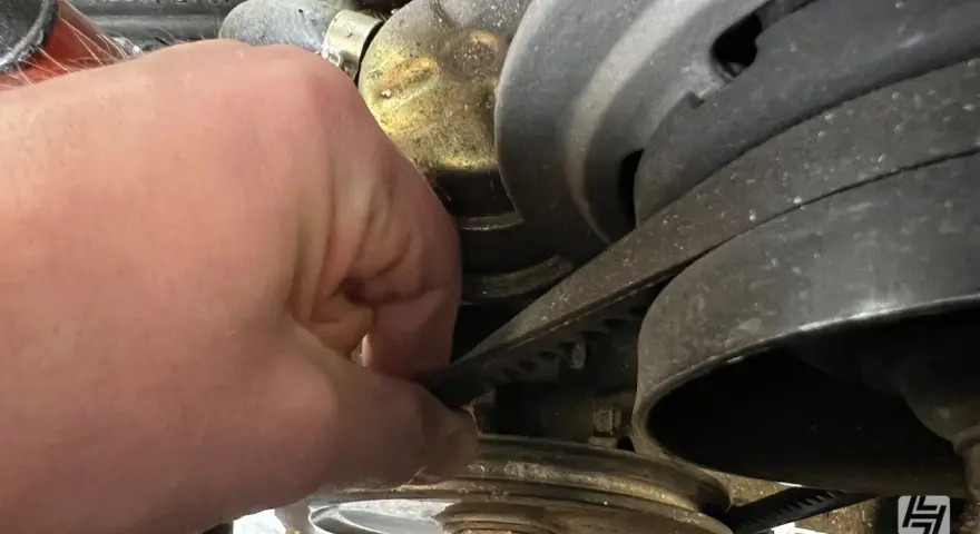 Hand prüft Keilriemen, der beim Anfahren quietscht. Der Riemen läuft über mehrere Riemenscheiben im Motorraum.