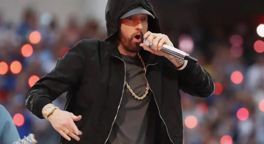 Eminem w czarnej bluzie z kapturem śpiewa do mikrofonu, w złotym łańcuchu i zegarku.